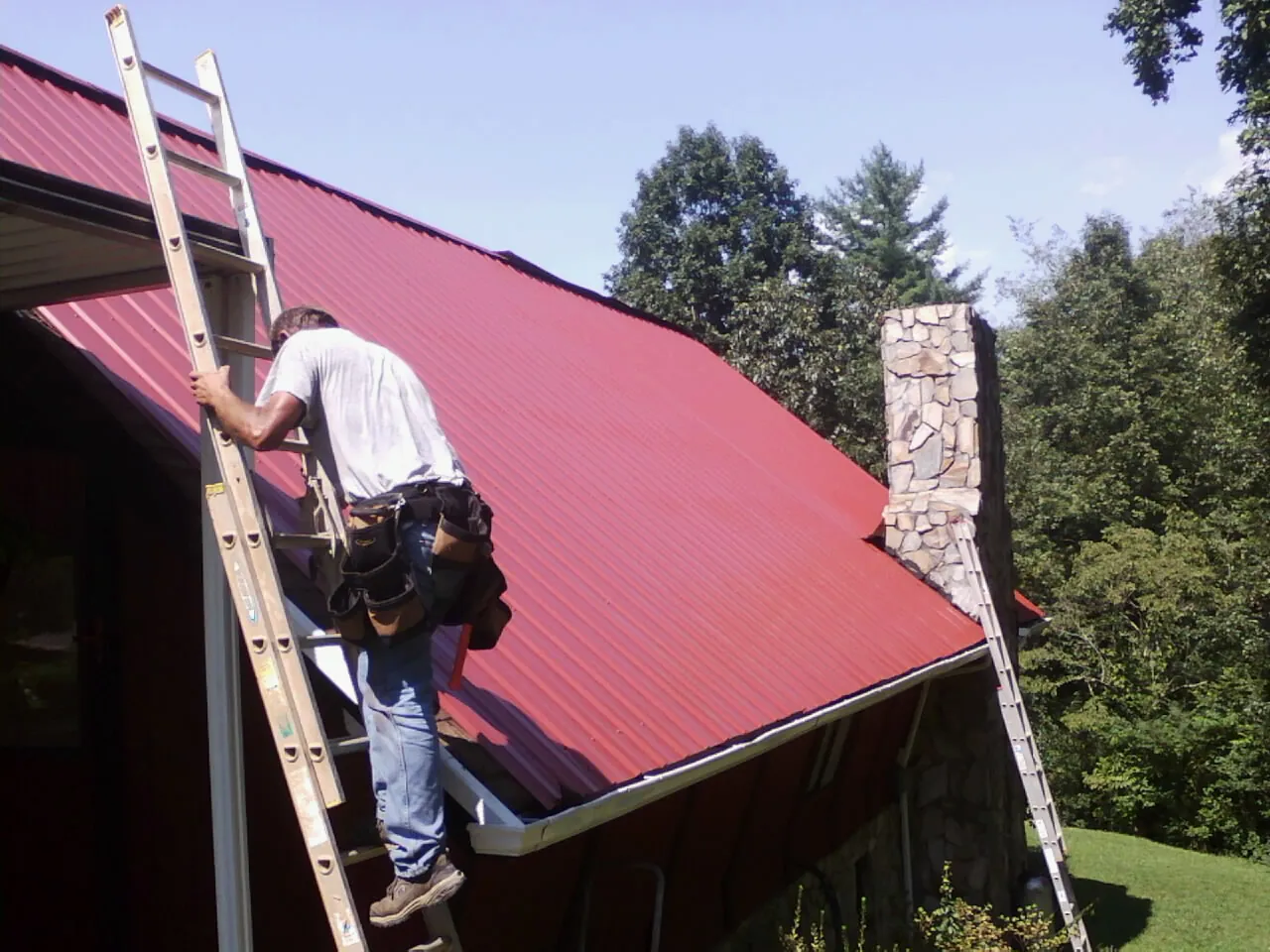 Roofer on a ladder beside a red metal roof and stone chimney — Soffit & Fascia Repair by Roofing Brothers in Nixa